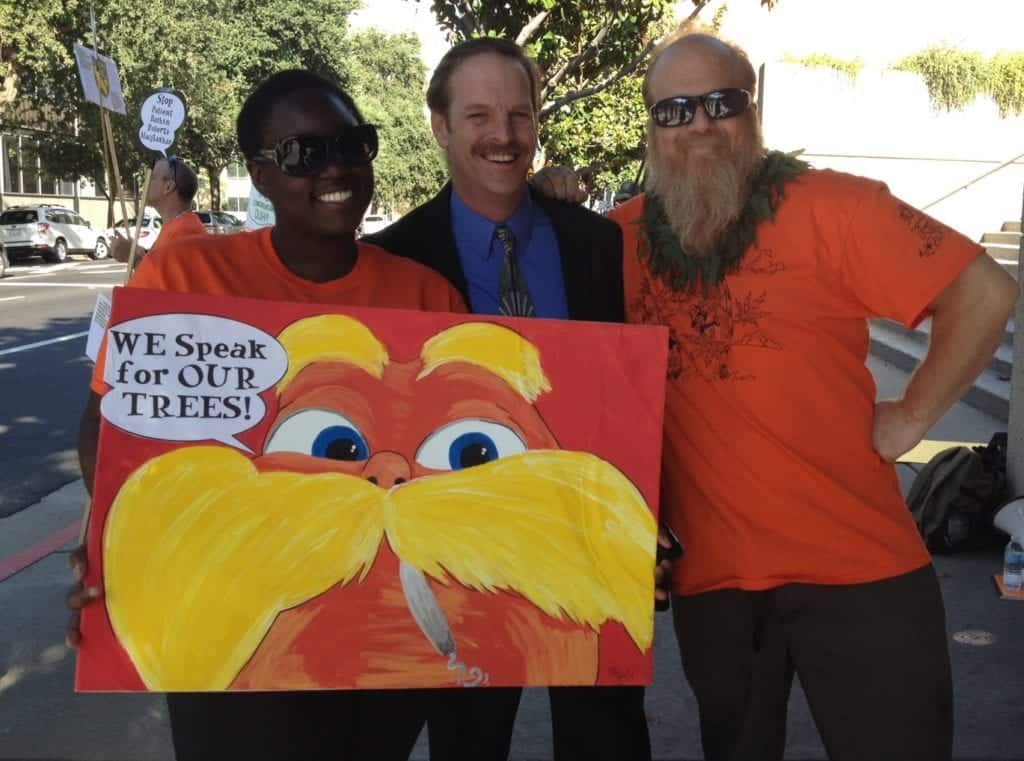 Three people stand outside; the person on the left holds a colorful sign with a character featuring a big orange mustache and the words "WE Speak for OUR TREES!" The other two, wearing Sacramento Supes shirts, smile at the camera. Trees and a sidewalk are visible in the background. CA Norml