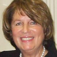 A smiling woman with short, light brown hair. She is wearing a dark top and a single strand pearl necklace, her confident demeanor reminiscent of a poised government witness. The background appears to be indoors with light-colored walls. CA Norml