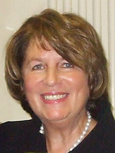 A smiling woman with short, light brown hair. She is wearing a dark top and a single strand pearl necklace, her confident demeanor reminiscent of a poised government witness. The background appears to be indoors with light-colored walls. CA Norml