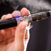 A close-up of a person vaping with an electronic cigarette. The individual's hand is holding the black and silver e-cigarette, which emits vapor, and the mouthpiece is near their lips. A blue light is illuminated on the device. Public housing policies often discourage such activities. The background is blurred. CA Norml