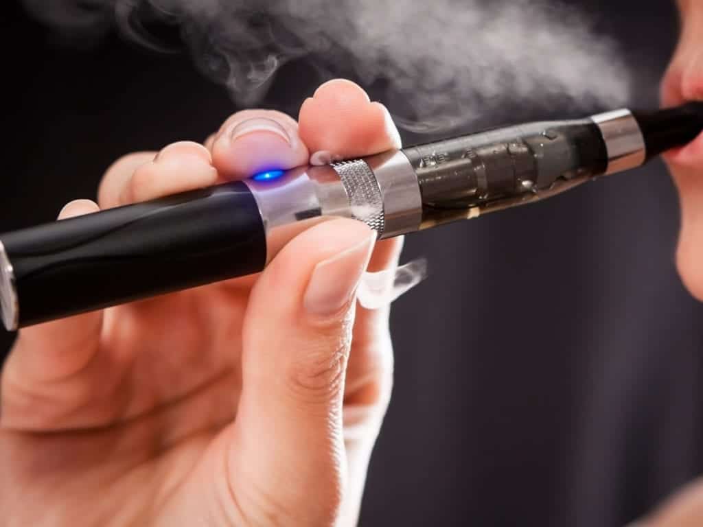 A close-up of a person vaping with an electronic cigarette. The individual's hand is holding the black and silver e-cigarette, which emits vapor, and the mouthpiece is near their lips. A blue light is illuminated on the device. Public housing policies often discourage such activities. The background is blurred. CA Norml