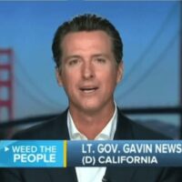 A man in a suit and white shirt speaks in front of an image of the Golden Gate Bridge. The text on the screen reads, "WEED THE PEOPLE" and "LT. GOV. GAVIN NEWSOM (D) CALIFORNIA." Amid growing concerns over Jeff Sessions' stance on marijuana prosecution, Newsom advocates for California's cannabis laws. CA Norml