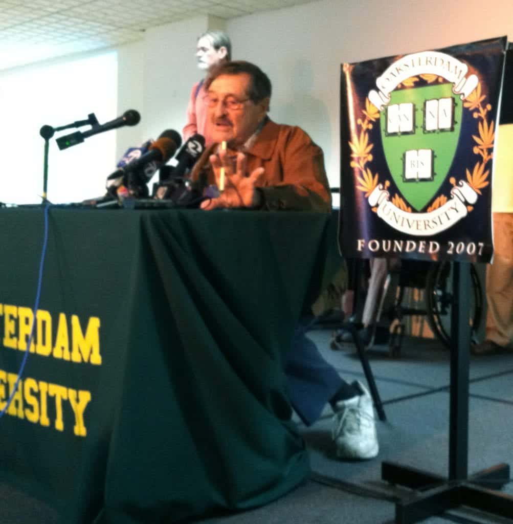 A person sits at a table adorned with a green cloth that reads "Amsterdam University" in yellow, speaking into one of the multiple microphones placed before them. Next to the person is a banner that says, "Amsterdam University Founded 2007." They mention John Vasconcellos and his contributions to human rights. CA Norml