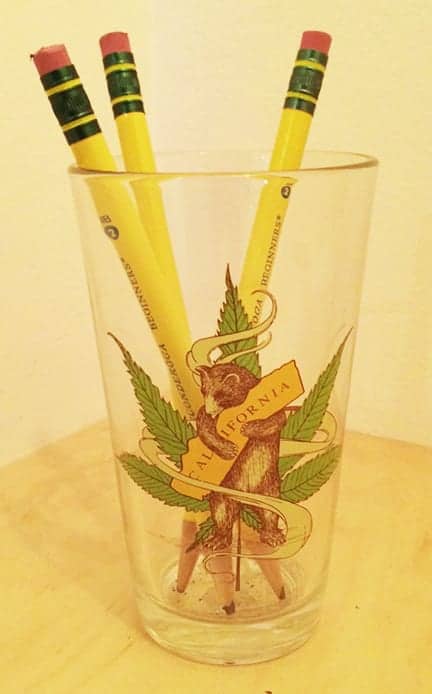 A clear glass with an illustration of a bear holding a banner that reads "California" and standing in front of a green cannabis leaf, nodding to CA Cannabis Regulations. The glass contains three yellow pencils with erasers and is placed on a wooden surface against a plain background. CA Norml