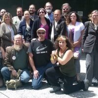 A group of about 20 people posing outside a building in Sacramento County. They are arranged in two rows, with some standing and others kneeling or sitting. Dressed in casual and business attire, they smile at the camera, seemingly unaffected by the outdoor grows ban currently in place. CA Norml