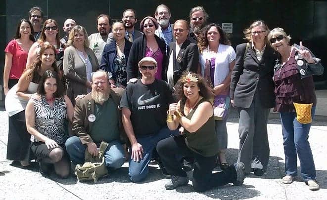 A group of about 20 people posing outside a building in Sacramento County. They are arranged in two rows, with some standing and others kneeling or sitting. Dressed in casual and business attire, they smile at the camera, seemingly unaffected by the outdoor grows ban currently in place. CA Norml