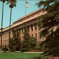 A grand, neoclassical building is pictured behind a well-manicured lawn adorned with tall palm trees and lush green bushes. Two flags flutter atop the building's rooftop, signaling local restrictions, while the sky is clear and blue. The architecture features columns and large windows. CA Norml