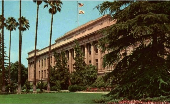 A grand, neoclassical building is pictured behind a well-manicured lawn adorned with tall palm trees and lush green bushes. Two flags flutter atop the building's rooftop, signaling local restrictions, while the sky is clear and blue. The architecture features columns and large windows. CA Norml