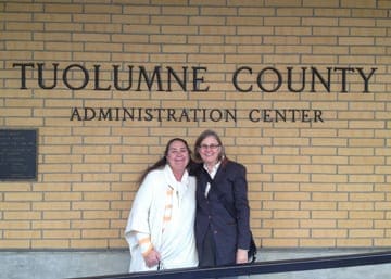 Two women standing and smiling in front of the Tuolumne County Administration Center. The woman on the left is wearing a white garment, and the woman on the right is wearing a dark jacket and glasses. They appear happy and are posing closely together, perhaps discussing the recent dispensary ban in Tuolumne County. CA Norml