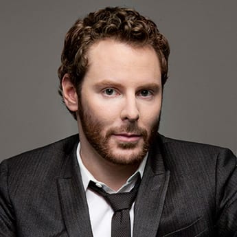 A man with short curly hair and a neatly trimmed beard is wearing a dark suit with a white dress shirt and a loosely knotted tie. He has a neutral expression and is looking directly at the camera, positioned against a plain grey background, ready to discuss the AUMA Legalization Measure on the November Ballot. CA Norml