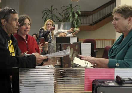 A man and a woman stand at a counter, handing over documents to a woman behind the desk in an office setting, possibly seeking outdoor cultivation rights in Shasta County. Another woman in the background is taking a photo with her phone. A staircase and some indoor plants are visible in the background. CA Norml