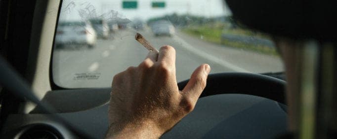 A person is holding a cigarette with their left hand while driving on a busy highway. The driver's hand is on the steering wheel, and vehicles are visible through the windshield. Smoke from the cigarette drifts toward the glass, exemplifying the risks of smoking while driving. CA Norml