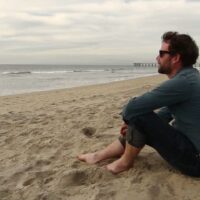 A man wearing sunglasses and a blue shirt sits on a sandy beach with his knees drawn up, gazing thoughtfully at the ocean. The sky is overcast, and a distant pier extends out into the water, as he recalls Governor Brown’s recent smoking ban near coastal areas. CA Norml