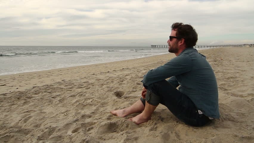 A man wearing sunglasses and a blue shirt sits on a sandy beach with his knees drawn up, gazing thoughtfully at the ocean. The sky is overcast, and a distant pier extends out into the water, as he recalls Governor Brown’s recent smoking ban near coastal areas. CA Norml