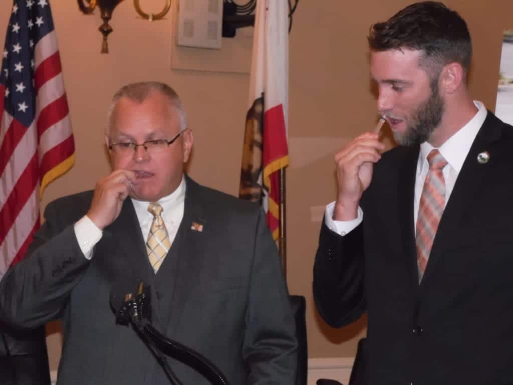 Two men in suits are standing indoors, holding up small objects to their mouths, possibly breathalyzers. The older man on the left is wearing glasses and a yellow tie, while the younger man on the right is wearing a black suit with a pink striped tie. Flags are visible in the background. CA Norml