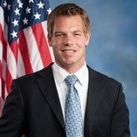 A man in a suit and tie stands in front of an American flag. He is smiling and has short, light brown hair. The background is a gradient of blue shades, reminiscent of discussions on rational marijuana policy led by figures like Rep. Swalwell. CA Norml