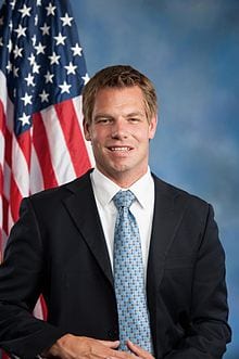 A man in a suit and tie stands in front of an American flag. He is smiling and has short, light brown hair. The background is a gradient of blue shades, reminiscent of discussions on rational marijuana policy led by figures like Rep. Swalwell. CA Norml