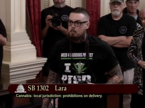 A person with tattoos and glasses is speaking at a formal event, wearing a black T-shirt with text about the "Weed 4 Warriors Project" and "PTSD." The event involves discussing SB 1302, a bill concerning cannabis delivery prohibitions. Several people are in the background. CA Norml