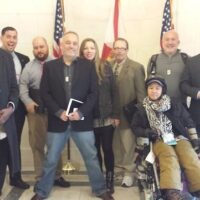 A group of nine people pose for a photo indoors, four women and five men, with an American flag and another flag in the background. One woman is in a wheelchair in the front row. The others, including several senators holding items like notebooks and ID badges, stand around her. CA Norml