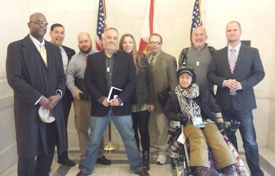 A group of nine people pose for a photo indoors, four women and five men, with an American flag and another flag in the background. One woman is in a wheelchair in the front row. The others, including several senators holding items like notebooks and ID badges, stand around her. CA Norml