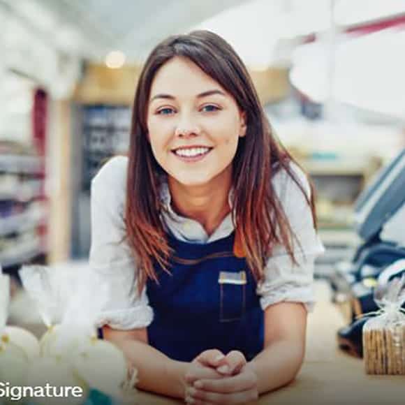 A person with long hair and a friendly smile is leaning on a counter, wearing a dark apron over a light shirt. Various packaged items are on the counter in the foreground. The background appears to be a store. CA Norml