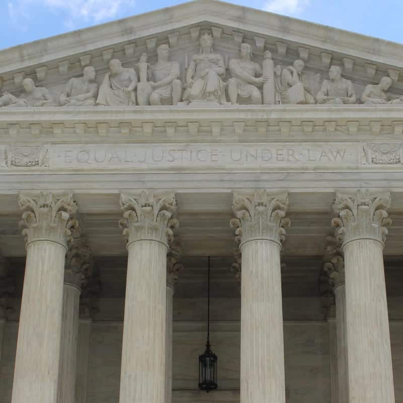 A close-up view of the U.S. Supreme Court building's facade, featuring intricate sculptures of figures and the engraved words "Equal Justice Under Law" above tall, fluted columns, under a pediment. A hanging lantern is seen between the columns. CA Norml