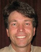 A man with short brown hair smiling at the camera. He is wearing a light-colored shirt, and there is a dark background behind him. CA Norml