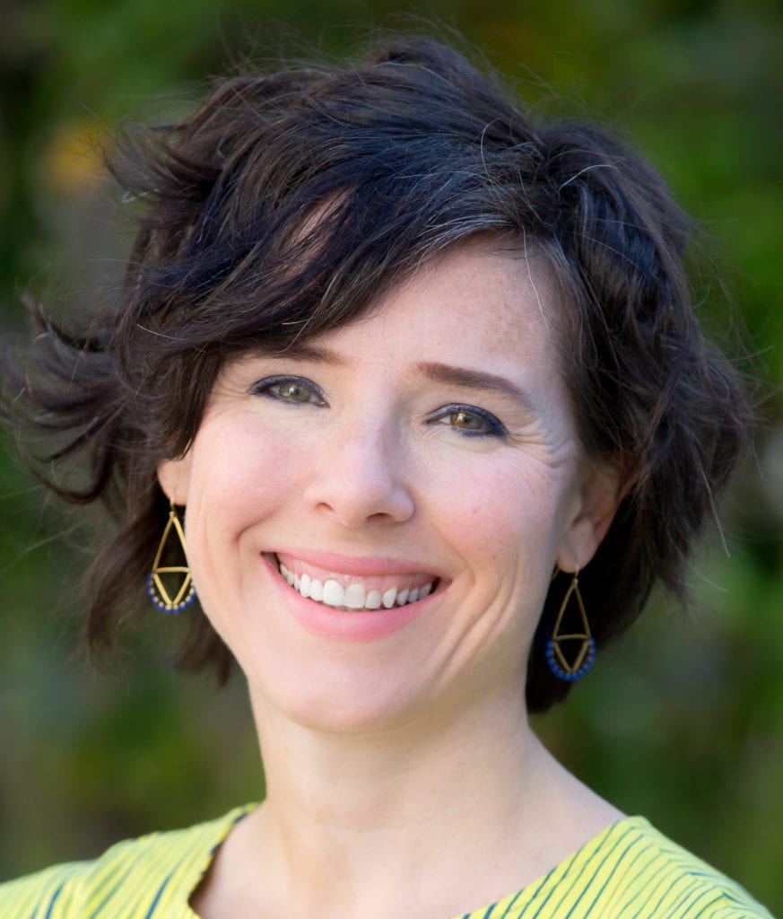 A woman with short, wavy dark hair and an asymmetrical part smiles warmly. She wears geometric earrings and a striped yellow top. The background is a blurred outdoor setting. CA Norml