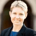 A person with short blonde hair is smiling while wearing a dark blazer and a blue top. The background is blurred, featuring an archway or similar structure. CA Norml