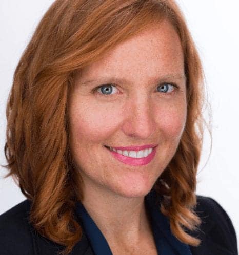 A woman with shoulder-length wavy auburn hair smiles at the camera. She is wearing a dark blazer over a dark blue top. The background is plain white. CA Norml