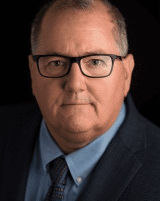 A man with short gray hair and glasses is wearing a blue dress shirt, patterned tie, and dark suit jacket. He is facing the camera with a neutral expression against a black background. CA Norml