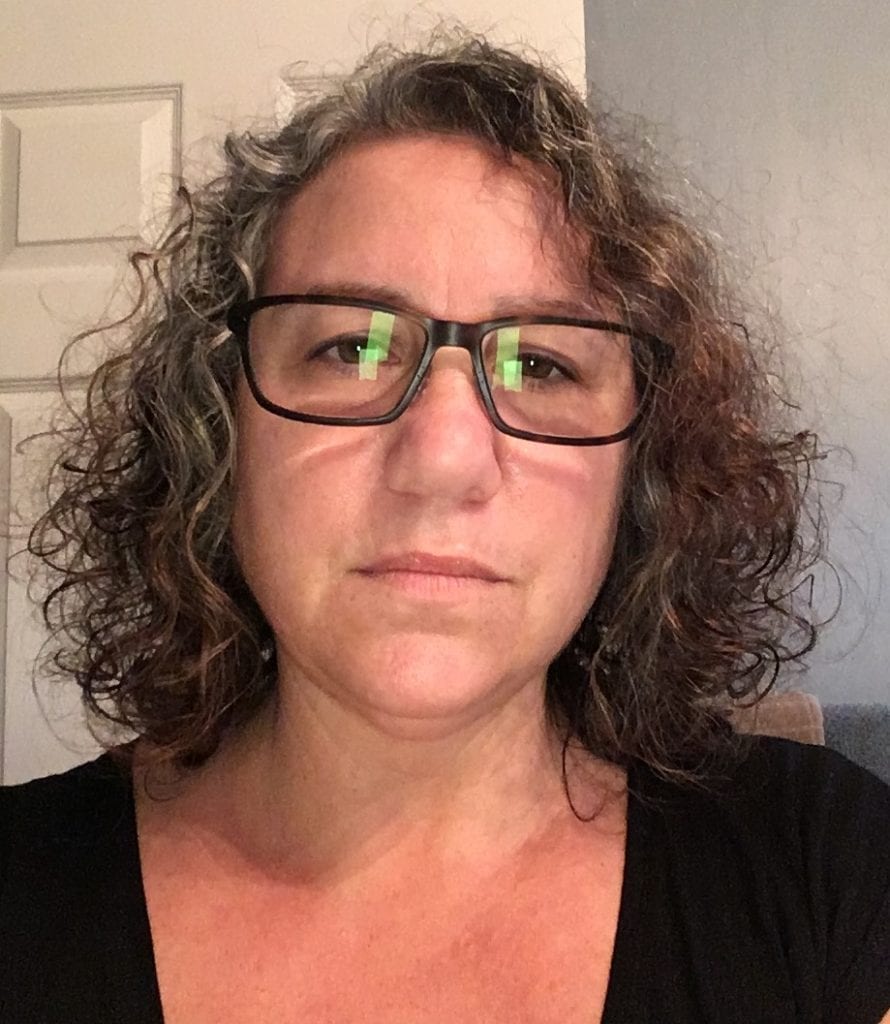 A person with short, curly hair, wearing glasses and a black shirt, is looking directly at the camera with a neutral expression. They are indoors with a door and a wall visible in the background—likely taken during the California NORML Legal Seminar in June 2021. CA Norml