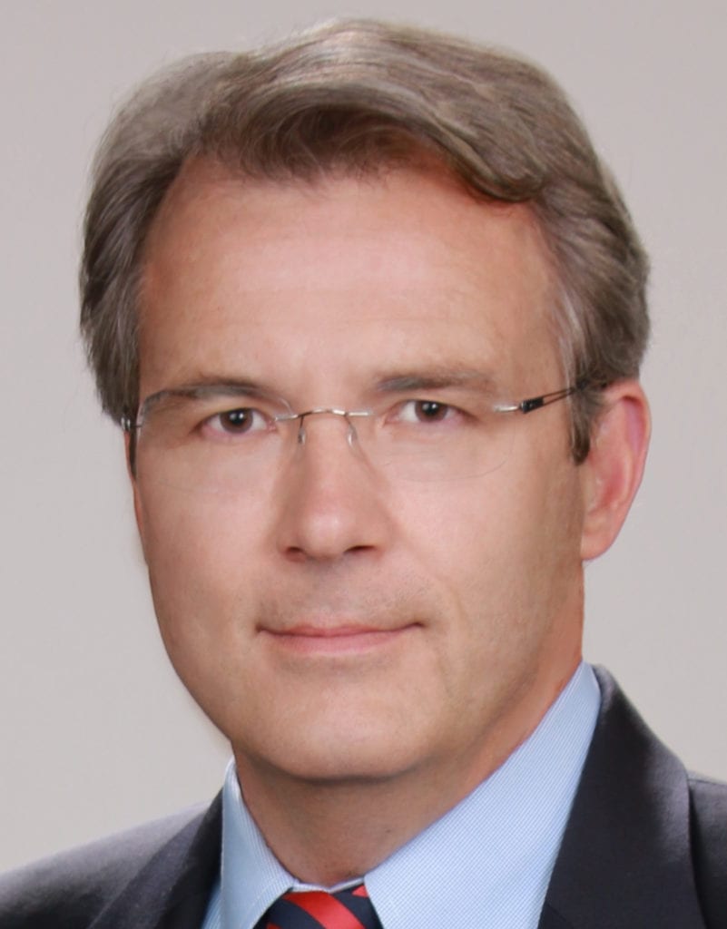 A middle-aged man with short, graying hair, wearing glasses and a business suit with a light blue shirt and red and blue striped tie, is looking directly at the camera against a plain background. CA Norml