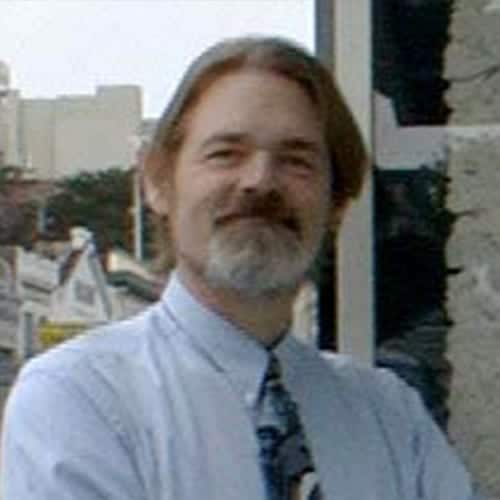 A middle-aged man with shoulder-length hair and a beard stands outside, smiling. He is wearing a light blue dress shirt and a patterned tie. Buildings and trees are visible in the background, creating an inviting scene for staff gatherings or team events. CA Norml