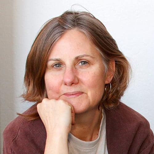 A middle-aged woman with short, light brown hair and blue eyes gazes at the camera. She rests her chin on her fist, wearing a neutral expression. Dressed in a light shirt with a brownish outer layer, she seems deep in thought. The plain white background adds to the focus on her contemplative demeanor, perhaps pondering about her team of employees. CA Norml