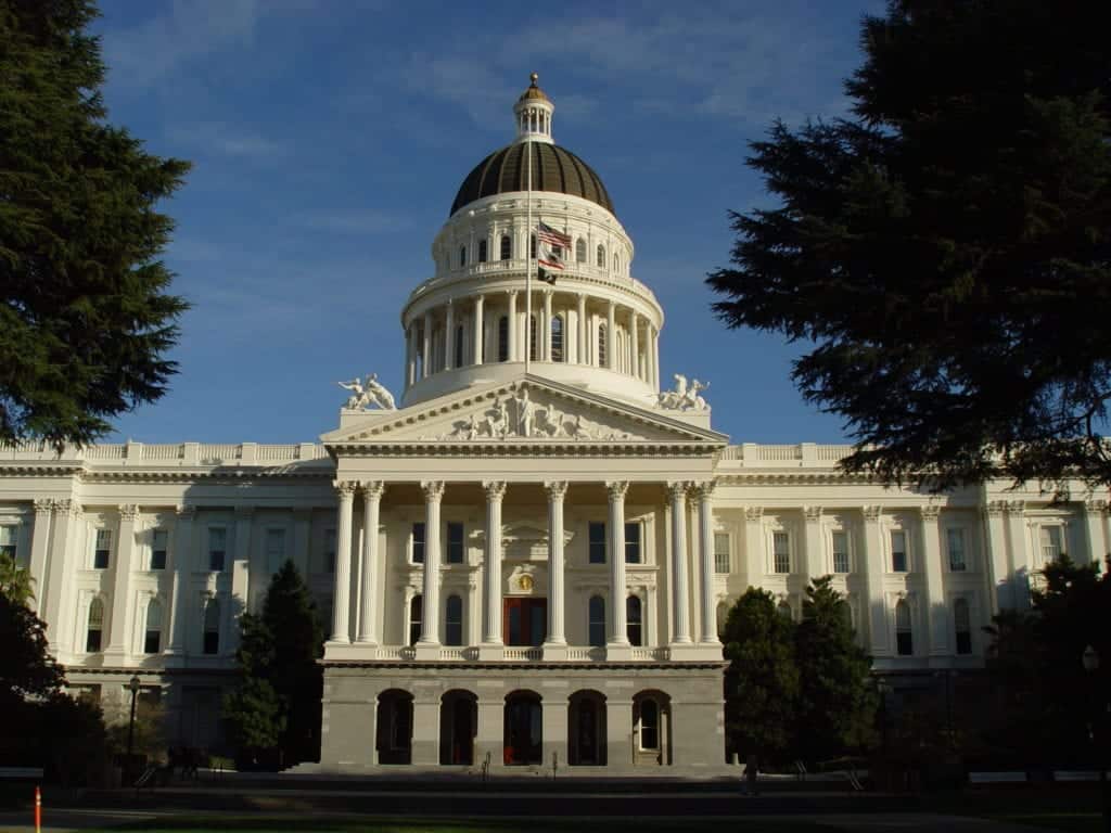 The image shows a large white classical-style building with a prominent dome, decorated with a small flag on top, and flanked by two wings. The building, where locals once banned cannabis deliveries, is surrounded by lush green trees and set against a clear blue sky. CA Norml