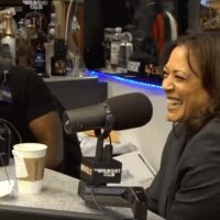 Two people are sitting at a radio studio table, laughing and engaged in conversation. The table has microphones, a laptop, and various beverages. The backdrop shows a variety of bottles. "The Breakfast Club" logo is visible in the bottom right corner as they discuss topics like marijuana legalization and Kamala Harris. CA Norml