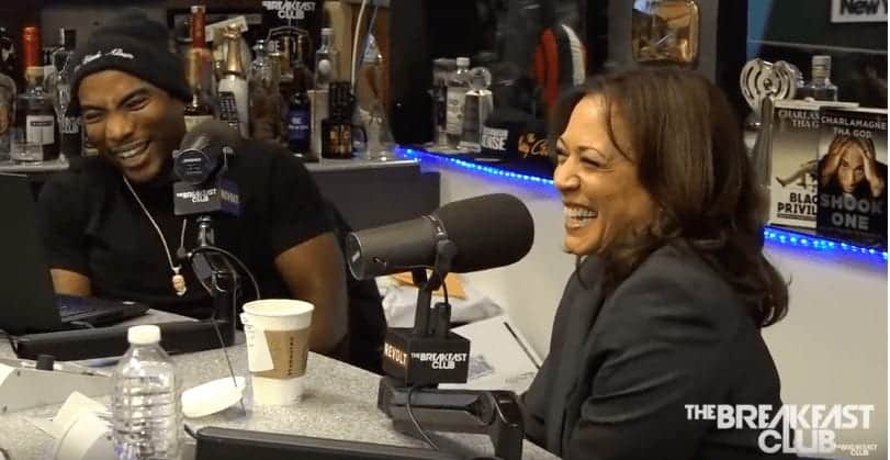 Two people are sitting at a radio studio table, laughing and engaged in conversation. The table has microphones, a laptop, and various beverages. The backdrop shows a variety of bottles. "The Breakfast Club" logo is visible in the bottom right corner as they discuss topics like marijuana legalization and Kamala Harris. CA Norml