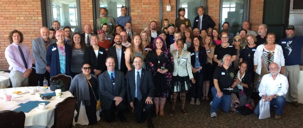 A diverse group of people pose for a group photo indoors in Sacramento, in front of a brick wall with large windows. Some are seated while most are standing. They are all smiling, and a few are holding awards or certificates from the Annual Lobby Day on May 6. Tables with place settings are visible in the foreground. CA Norml