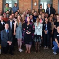 A large group of people standing and posing for a picture indoors. The individuals vary in age, gender, and attire. They seem to be gathered for a formal or semi-formal event, Annual Lobby Day in Sacramento, with some wearing name tags. The setting appears to be a spacious room with brick walls. CA Norml