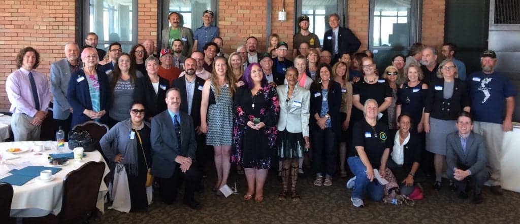 A large group of people standing and posing for a picture indoors. The individuals vary in age, gender, and attire. They seem to be gathered for a formal or semi-formal event, Annual Lobby Day in Sacramento, with some wearing name tags. The setting appears to be a spacious room with brick walls. CA Norml