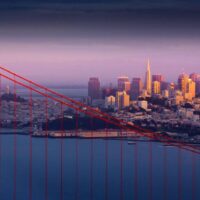 A scenic view of San Francisco at sunset with the Golden Gate Bridge in the foreground. The city's skyline, including high-rise buildings, is illuminated by a warm, golden light, while the sky is filled with a gradient from dark to light hues. San Francisco's progressive stance on marijuana convictions adds to its modern charm. CA Norml