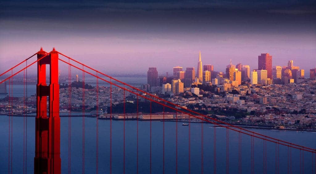 A scenic view of San Francisco at sunset with the Golden Gate Bridge in the foreground. The city's skyline, including high-rise buildings, is illuminated by a warm, golden light, while the sky is filled with a gradient from dark to light hues. San Francisco's progressive stance on marijuana convictions adds to its modern charm. CA Norml