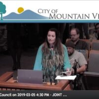 A woman stands at a podium addressing the city council of Mountain View about ordinance changes. She has long hair and is wearing a blue and brown outfit. Seated individuals in the background listen attentively. The display shows "City Council on 2019-03-05 4:30 PM - JOINT... CA Norml