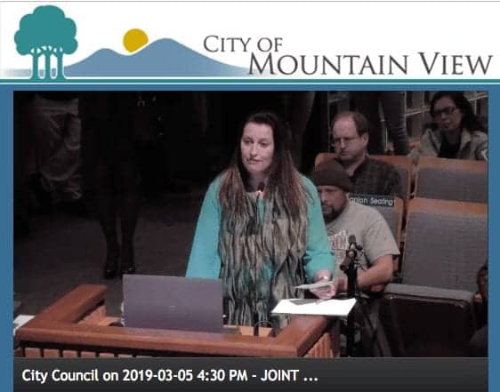 A woman stands at a podium addressing the city council of Mountain View about ordinance changes. She has long hair and is wearing a blue and brown outfit. Seated individuals in the background listen attentively. The display shows "City Council on 2019-03-05 4:30 PM - JOINT... CA Norml