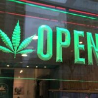 A bright green sign with a cannabis leaf and the word "OPEN" is displayed in the window of a store. The background shows the interior of the store with various items and posters promoting regulated cannabis, indicative of its presence in local markets. CA Norml