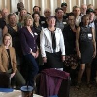 A large group of people in formal attire pose for a photo in a well-lit room with round tables and striped curtains in the background. There is a mix of males and females of various ages, some wearing name tags, gathered for Lobby Day organized by Cal NORML in Sacramento. CA Norml