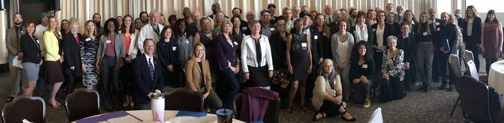 A large group of people in formal attire pose for a photo in a well-lit room with round tables and striped curtains in the background. There is a mix of males and females of various ages, some wearing name tags, gathered for Lobby Day organized by Cal NORML in Sacramento. CA Norml