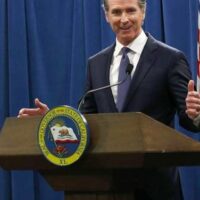 A man stands at a podium with a seal that says "Governor of the State of California." He is wearing a dark suit and tie, speaking into a microphone with his hands gesturing. Discussing statutory changes, the background is a blue curtain, and an American flag is partially visible on the side. CA Norml