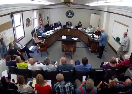 A large group of people are seated in a meeting room, facing a panel of individuals at a long table. The panel appears to be conducting a formal meeting regarding the ban on hemp cultivation in Plumas County. An American flag is visible in the corner of the room, and papers and documents are spread out in front of the participants. CA Norml